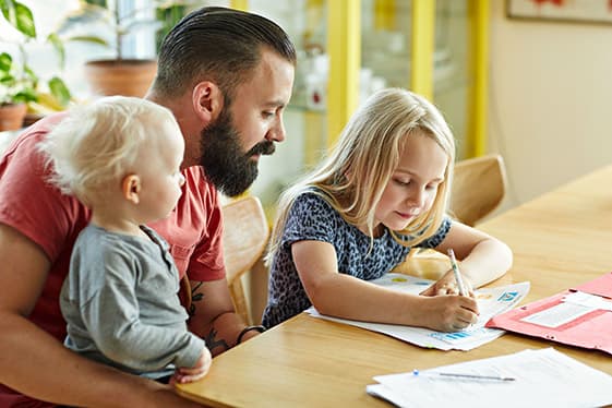 Pappa hjälper en yngre dotter med läxa vid köksbodet. Han har ett mindre barn i knät.