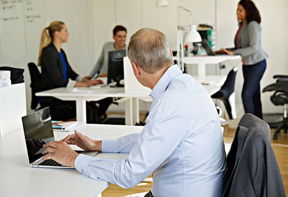 Man sitter vid en dator i ett kontorslandskap och vänder sig mot kollegor som diskuterar i bakgrunden