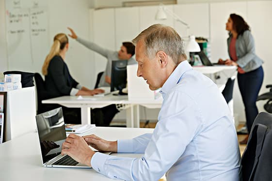 Man arbetar vid en dator i ett kontorslandskap och kollegor diskuterar i bakgrunden